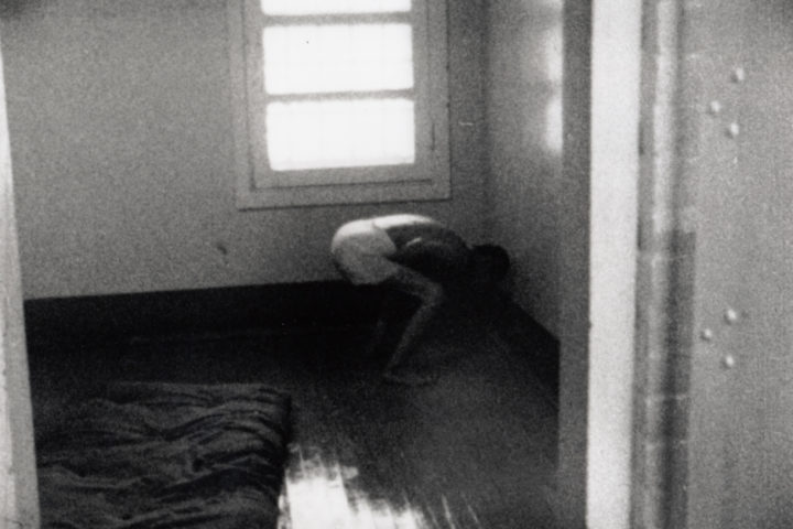 A black and white photo of a man in a barren room. He is wearing white boxer shorts and is bent over, screaming.
