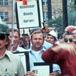 A group of protesters marches together. There are many men holding signs. At the front is a man in a red jacket and light pink shirt, and one with a tan shirt, green ballcap, and sunglasses.