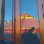 ] In a reflection of an unmarked storefront is a grayish silhouette of a man using an electric wheelchair. Behind the man is a spectacular red and yellow circus tent.