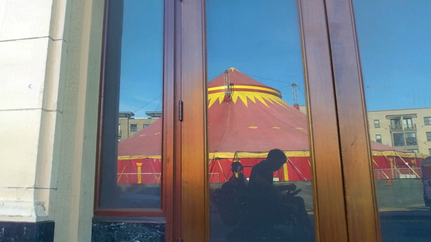 ] In a reflection of an unmarked storefront is a grayish silhouette of a man using an electric wheelchair. Behind the man is a spectacular red and yellow circus tent.