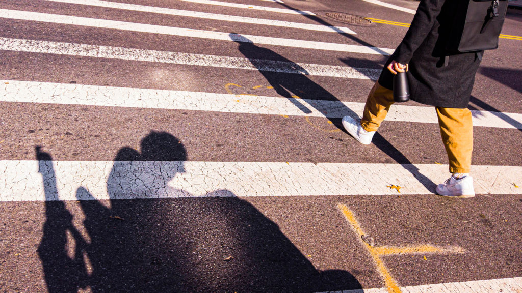 The shadow of a man in an electric wheelchair appears atop the white stripes of a city crosswalk. A woman in a black dress is walking through the intersection.