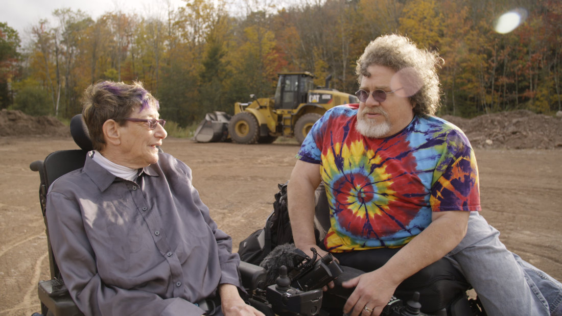 A man in a tie-dye shirt speaks with a woman in a grey collard shirt. They are both in wheelchairs.