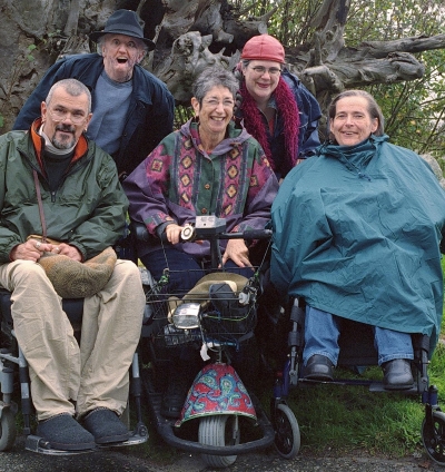 A row of three people in wheelchairs sit smiling in front of two friends who are standing