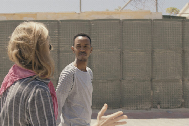 Michelle Shepard is a white woman with blond hair. She is wearing a purple plaid shirt with a pink scarf. She is facing Ismael Abdulle, a young Black man with black hair who is wearing a white long-sleeved shirt. They are standing in front of a barricade in Mogadishu.