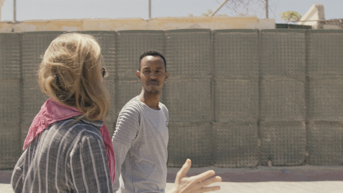 Michelle Shepard is a white woman with blond hair. She is wearing a purple plaid shirt with a pink scarf. She is facing Ismael Abdulle, a young Black man with black hair who is wearing a white long-sleeved shirt. They are standing in front of a barricade in Mogadishu.