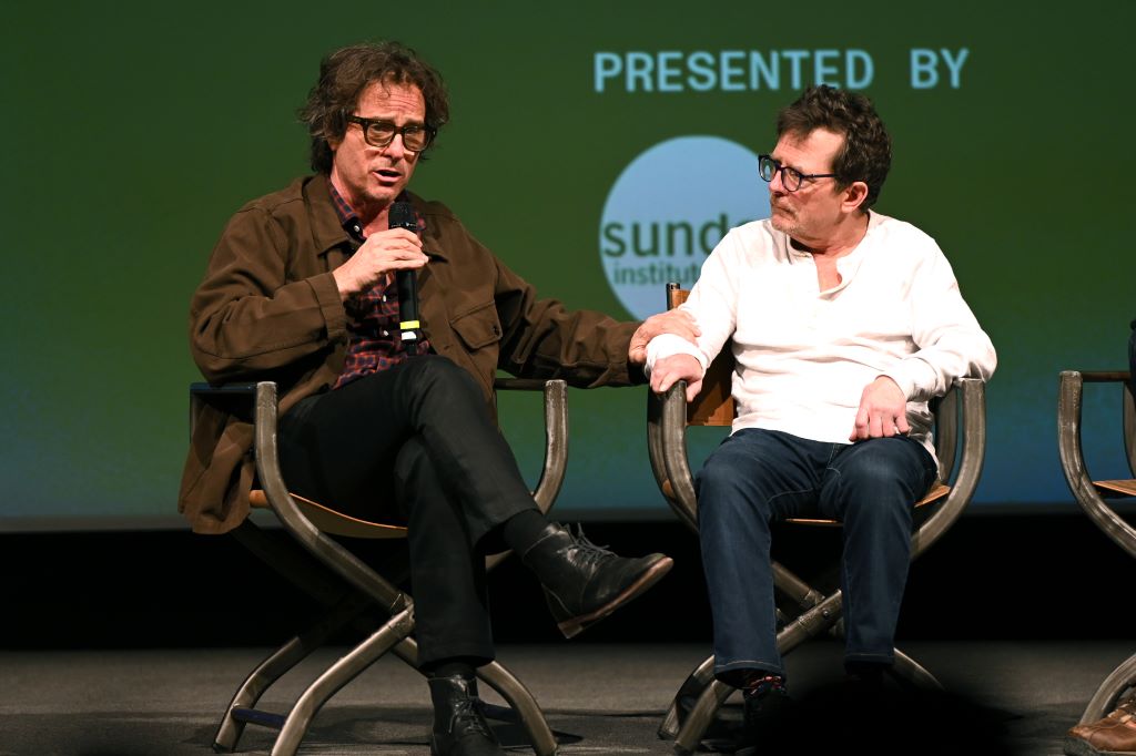 Director Davis Guggenheim and actor Michael J. Fox are seated onstage facing an audience. Guggenheim is wearing a brown jacket and black pants and is holding a microphone. Fox is wearing a white shirt and blue jeans.