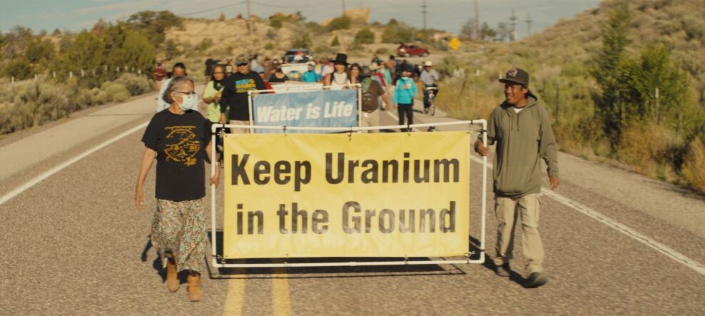 A group of Indigenous protesters marches on a desert road. They are carrying a yellow sign with black text that reads, "Keep uranium in the ground."