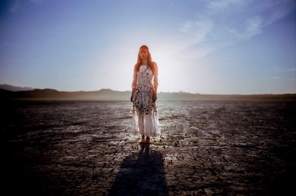 A young Indigenous woman stands in the desert. She is wearing a white dress and is standing in front of the sun with the light coming from behind her.