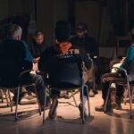 A film still from We Will Be Brave showing eight Good Guise members sitting together in a circle during one of their community sessions. It looks similar to a circle therapy session.