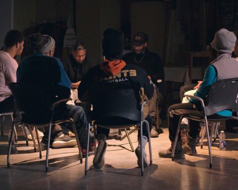 A film still from We Will Be Brave showing eight Good Guise members sitting together in a circle during one of their community sessions. It looks similar to a circle therapy session.