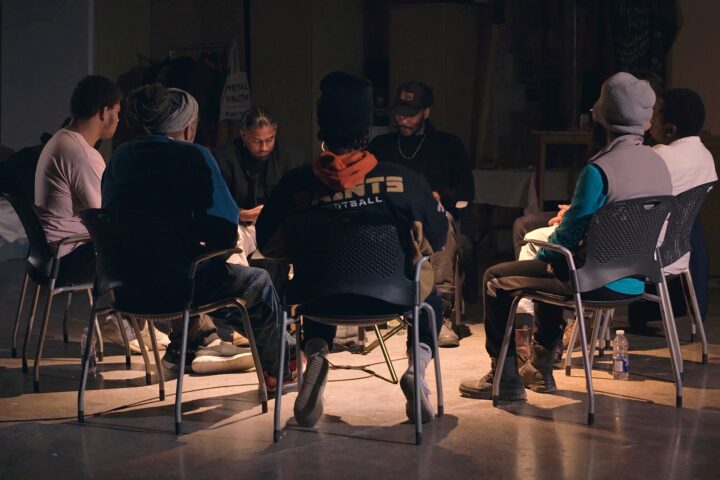 A film still from We Will Be Brave showing eight Good Guise members sitting together in a circle during one of their community sessions. It looks similar to a circle therapy session.