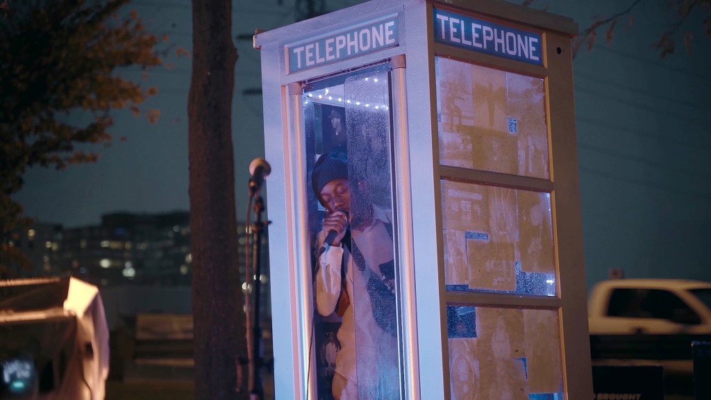 A film still from We Will Be Brave showing Beerus singing into a mic while inside a telephone booth.