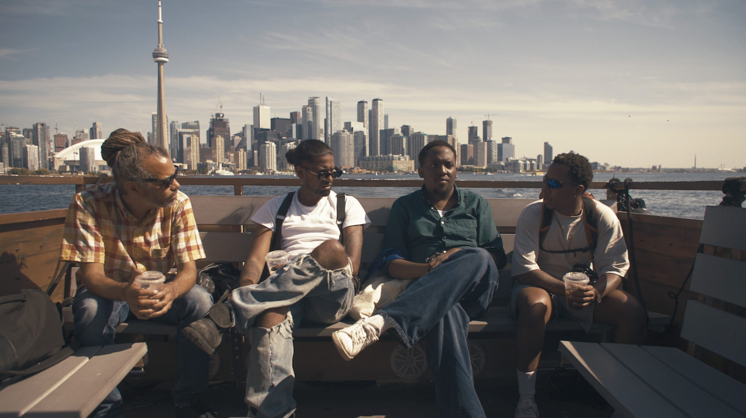 A film still from We Will Be Brave showing Julian, Balu, Bo and Jah Grey sitting on a water taxi, with the Toronto skyline in the background. 