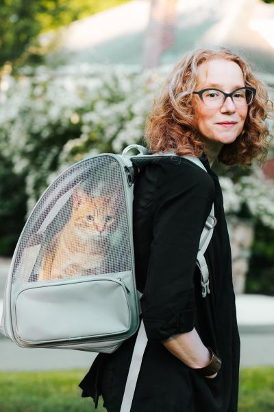 A woman with red hair and glasses stands posed with an orange cat in a backpack