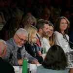 A diverse group of professionals at an industry conference sits at a board table. They are having an engaged conversation.