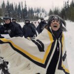 Witsuswit'en land defender Tsakë ze’ Howilhkat Freda Huson, stands in ceremony while police arrive to enforce Coastal GasLink’s injunction. She is wearing a traditional dress that is white with yellow and black markings.