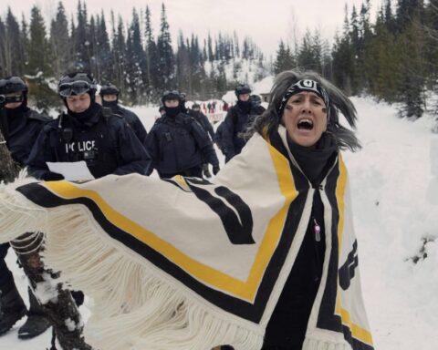Witsuswit'en land defender Tsakë ze’ Howilhkat Freda Huson, stands in ceremony while police arrive to enforce Coastal GasLink’s injunction. She is wearing a traditional dress that is white with yellow and black markings.