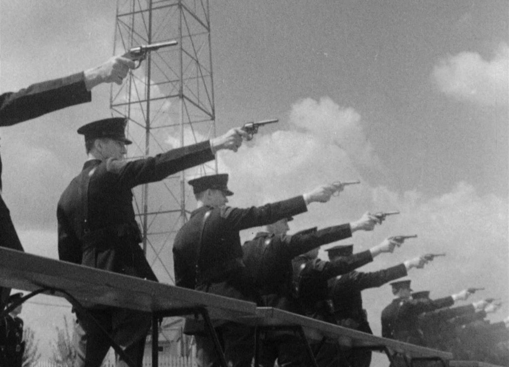 a black and white image of police officers in a row, they are firing guns and framed as if they are giving a Nazi salute.