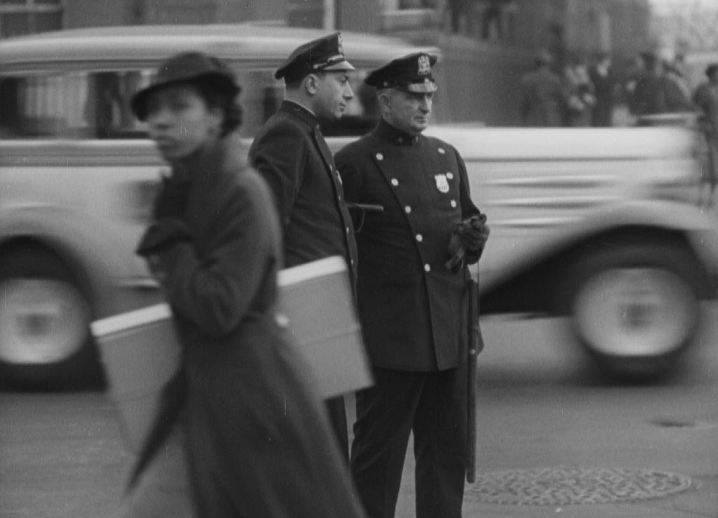 A black and white archival photo of a Black woman in an overcoat and hat walking quickly past two white police officers in uniform.
