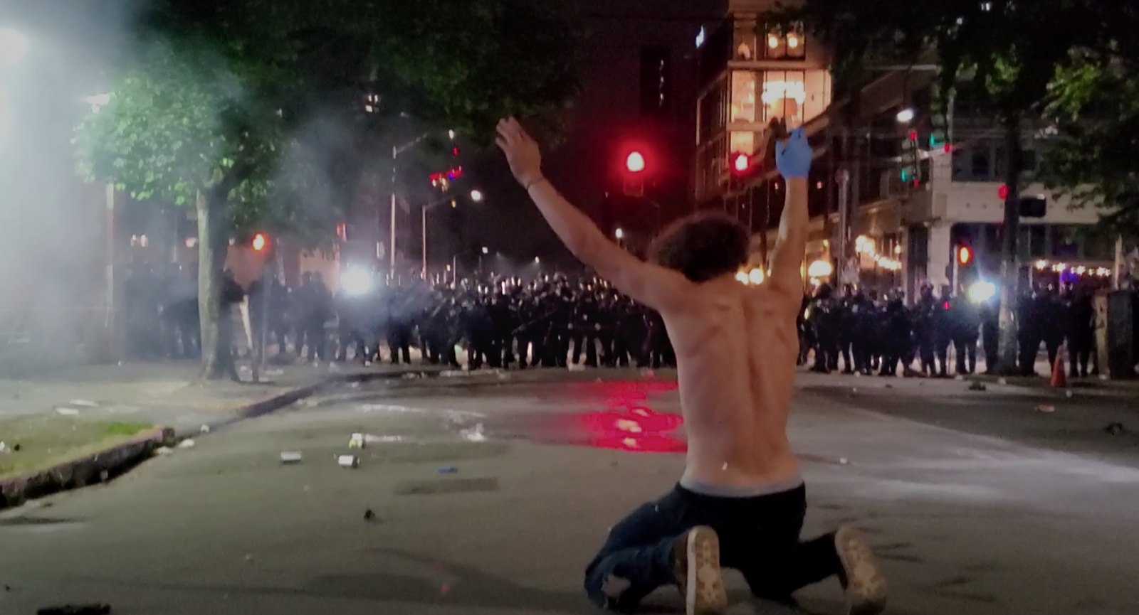 A protester kneels before a barricade of police officers in riot gear.