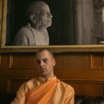 A Hare Krishna follower sits beneath a black and white portrait of his guru. The follower is a white man in an orange robe with a shaved head.