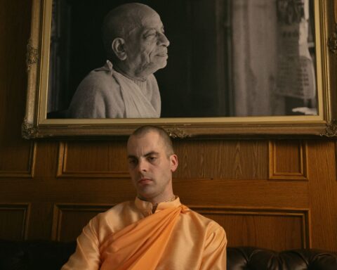 A Hare Krishna follower sits beneath a black and white portrait of his guru. The follower is a white man in an orange robe with a shaved head.