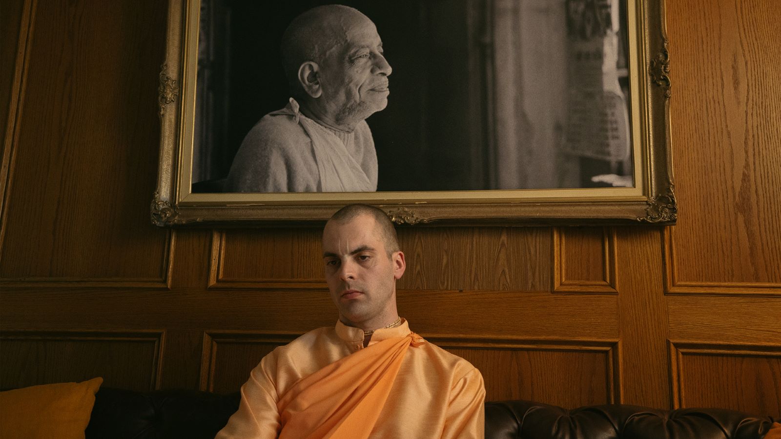 A Hare Krishna follower sits beneath a black and white portrait of his guru. The follower is a white man in an orange robe with a shaved head.