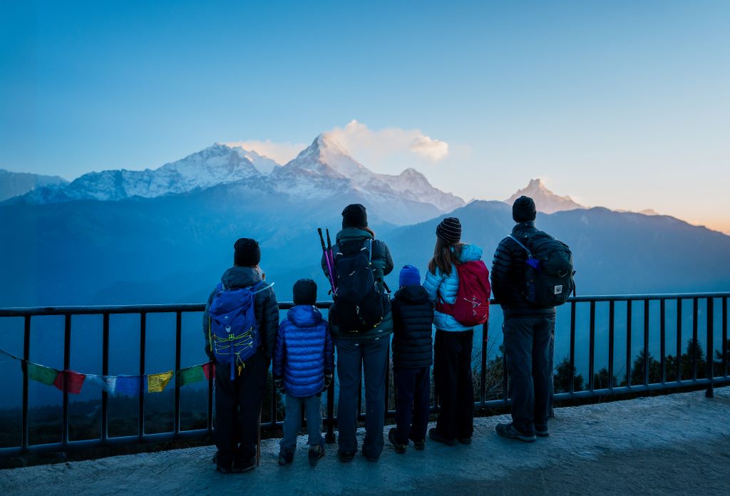 A family of two adults and four children look at a mountain range in Nepal. Their backs are to the camera and they are wearing snowsuits.