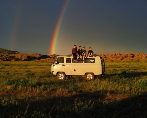 Four children sit on the roof of a white camper van in a field. There is a rainbow behind them across a dark blue sky.