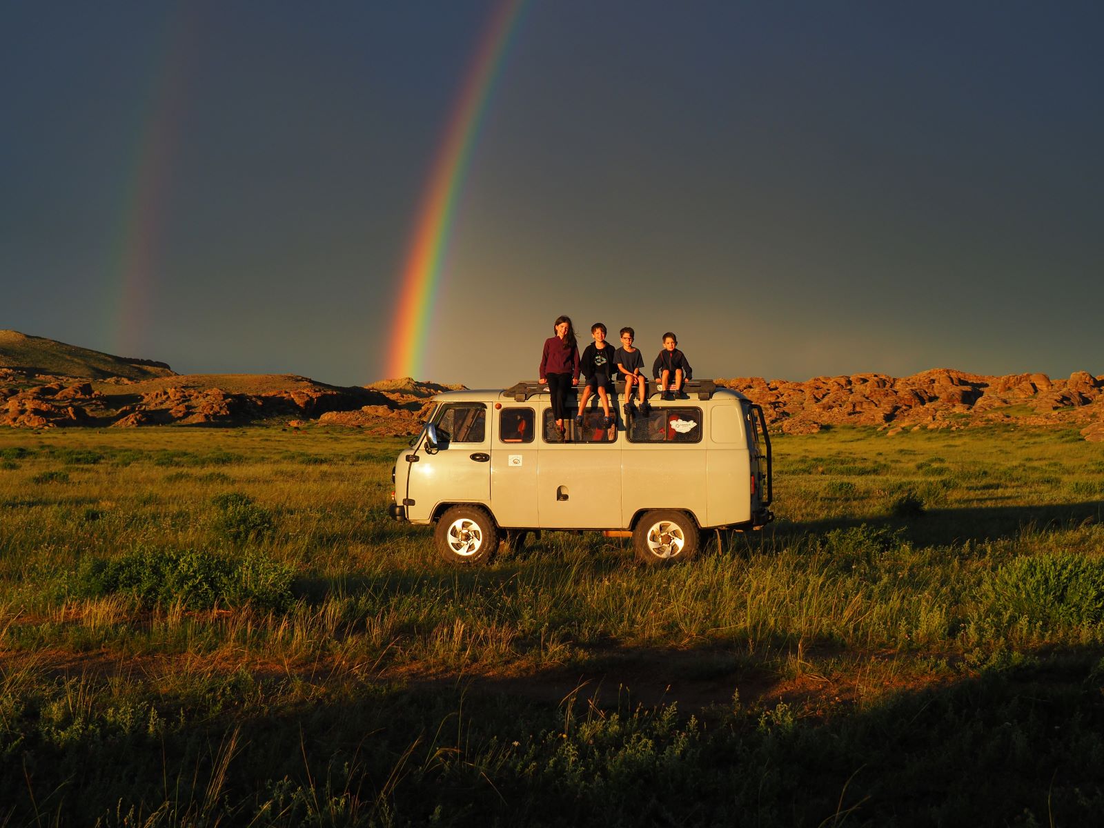 Four children sit on the roof of a white camper van in a field. There is a rainbow behind them across a dark blue sky.