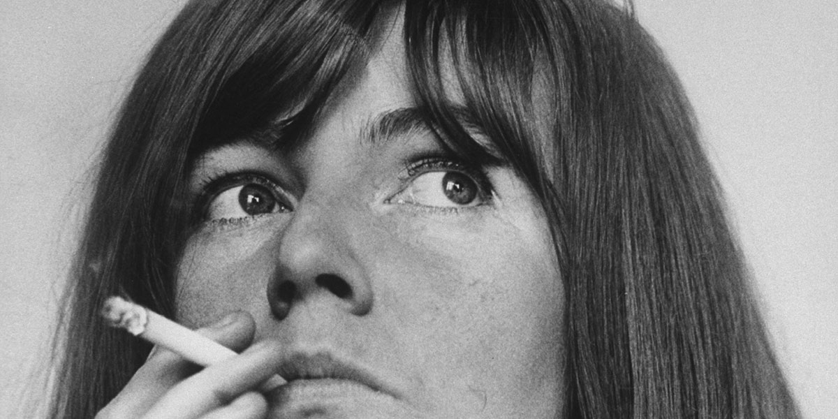 A black and white portrait of Irish author Edna O'Brien. She is framed in close-up while smoking a cigarette.