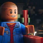 Singer/rapper Pharrell Williams as a LEGO man. He is sitting in a chair wearing a blue jacket with a red shirt.