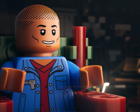 Singer/rapper Pharrell Williams as a LEGO man. He is sitting in a chair wearing a blue jacket with a red shirt.