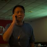 A Taiwanese man in his 70s speaks on a mobile telephone in a darkly-lit room. He is wearing a blue shirt and the TV is on.