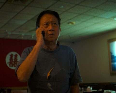 A Taiwanese man in his 70s speaks on a mobile telephone in a darkly-lit room. He is wearing a blue shirt and the TV is on.