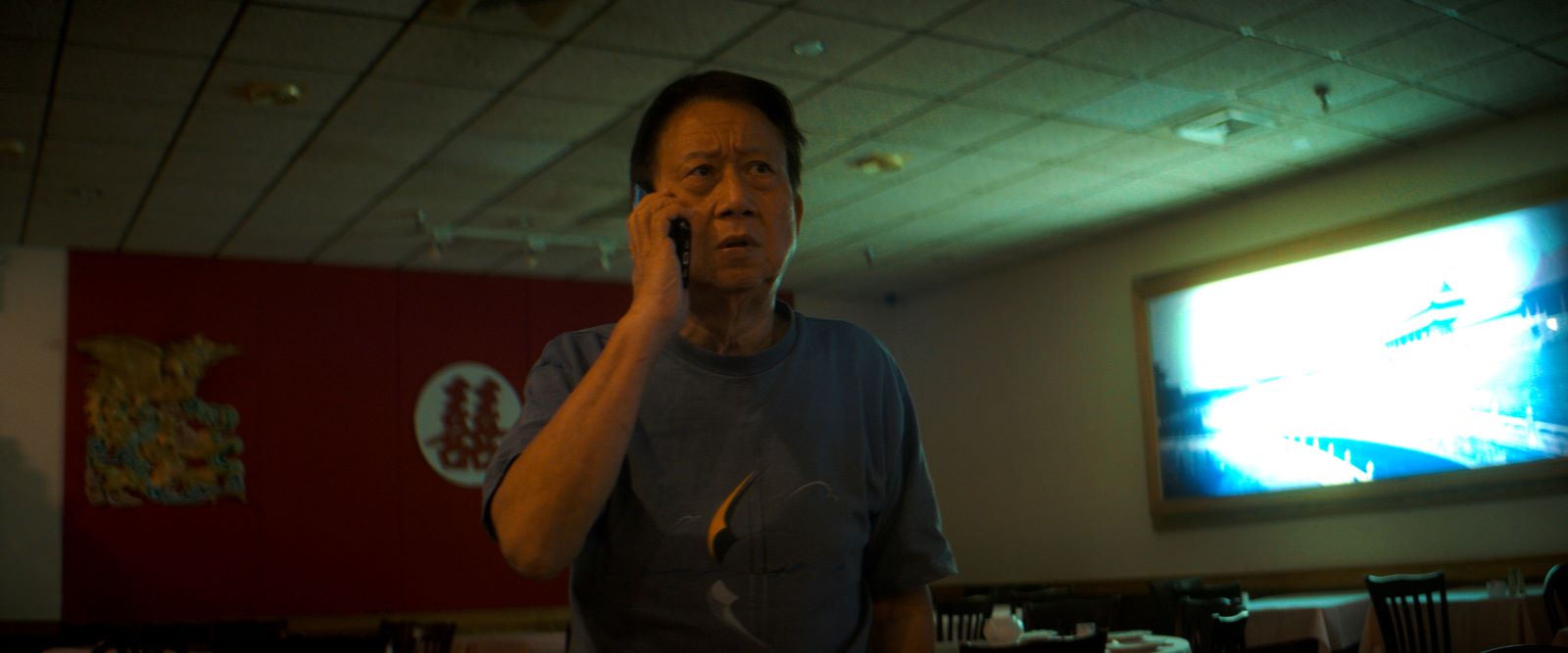 A Taiwanese man in his 70s speaks on a mobile telephone in a darkly-lit room. He is wearing a blue shirt and the TV is on.