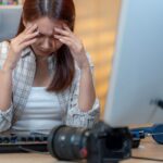 A woman sits at her computer with her hands on her forehead, indicating that she had a headache.