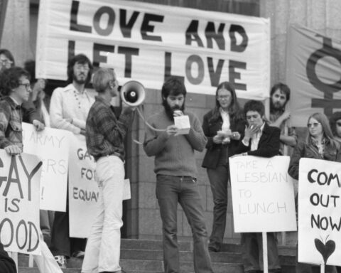 A Black and white archival photo of LGBTQ+ activists staging a protest with a megaphone and signs saying 'Love and Let Love.'