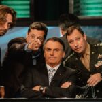 Former Brazilian president Jair Bolsonaro sits at a desk with his arms folded defiantly. He is surrounded by four men in suits. They are all looking at the camera.