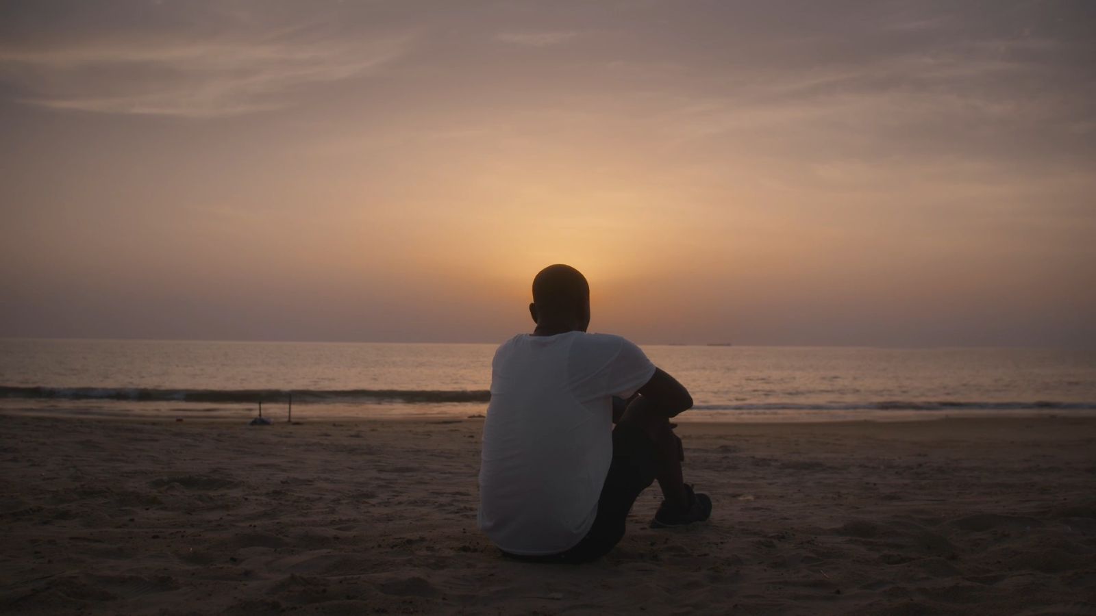 A man sits facing the sunset. His back is to the camera and he is wearing a white shirt.
