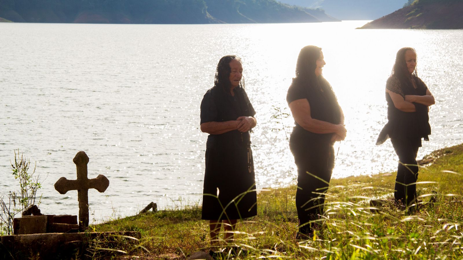 Three women sing by the water. They are wearing black veils and there is a cross on their left.