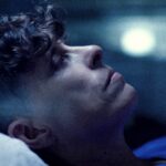 A close up of poet Andrea Gibson. They are lying down looking forward reflectively. They are Caucasian with dark brown hair that is long at the top and shaved on the sides. The photo has a dark blue hue.