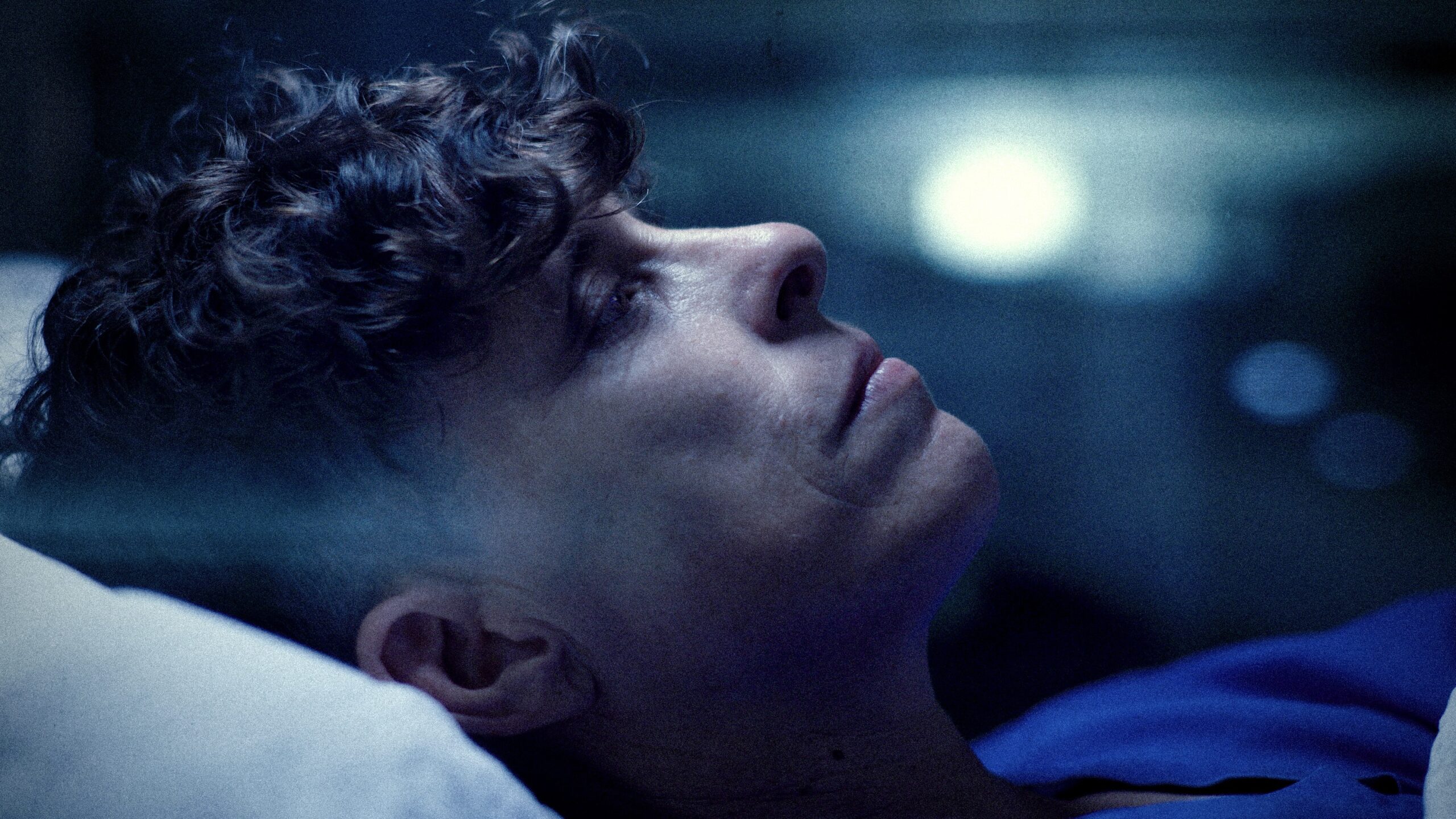 A close up of poet Andrea Gibson. They are lying down looking forward reflectively. They are Caucasian with dark brown hair that is long at the top and shaved on the sides. The photo has a dark blue hue.