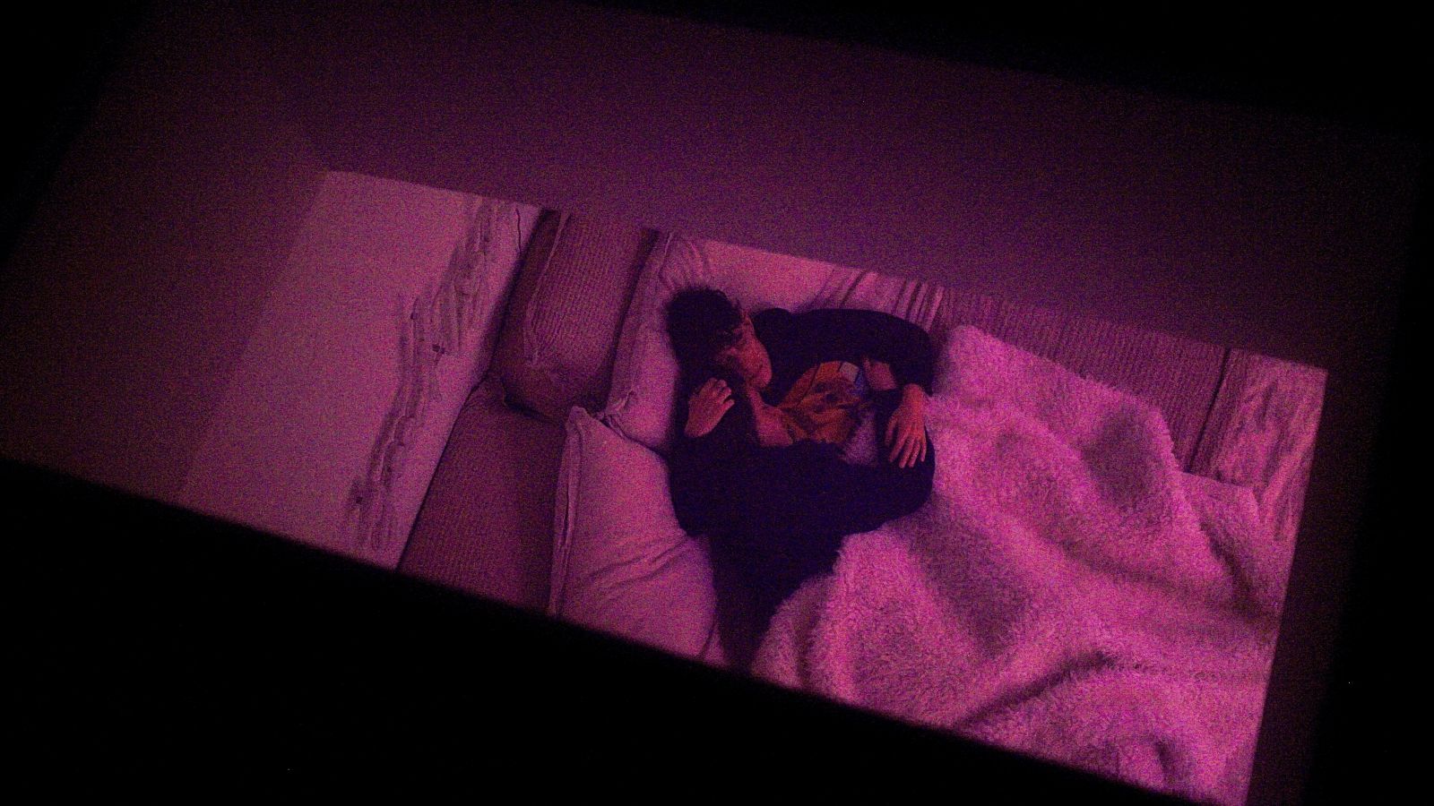 Two partners embrace in bed. They are viewed from above. Their bed has white sheets and the image has a pink hue.
