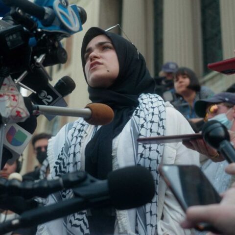 Maya Abdallah speaks into microphones during a press scrum. She is wearing a keffiyeh and a black hijab.