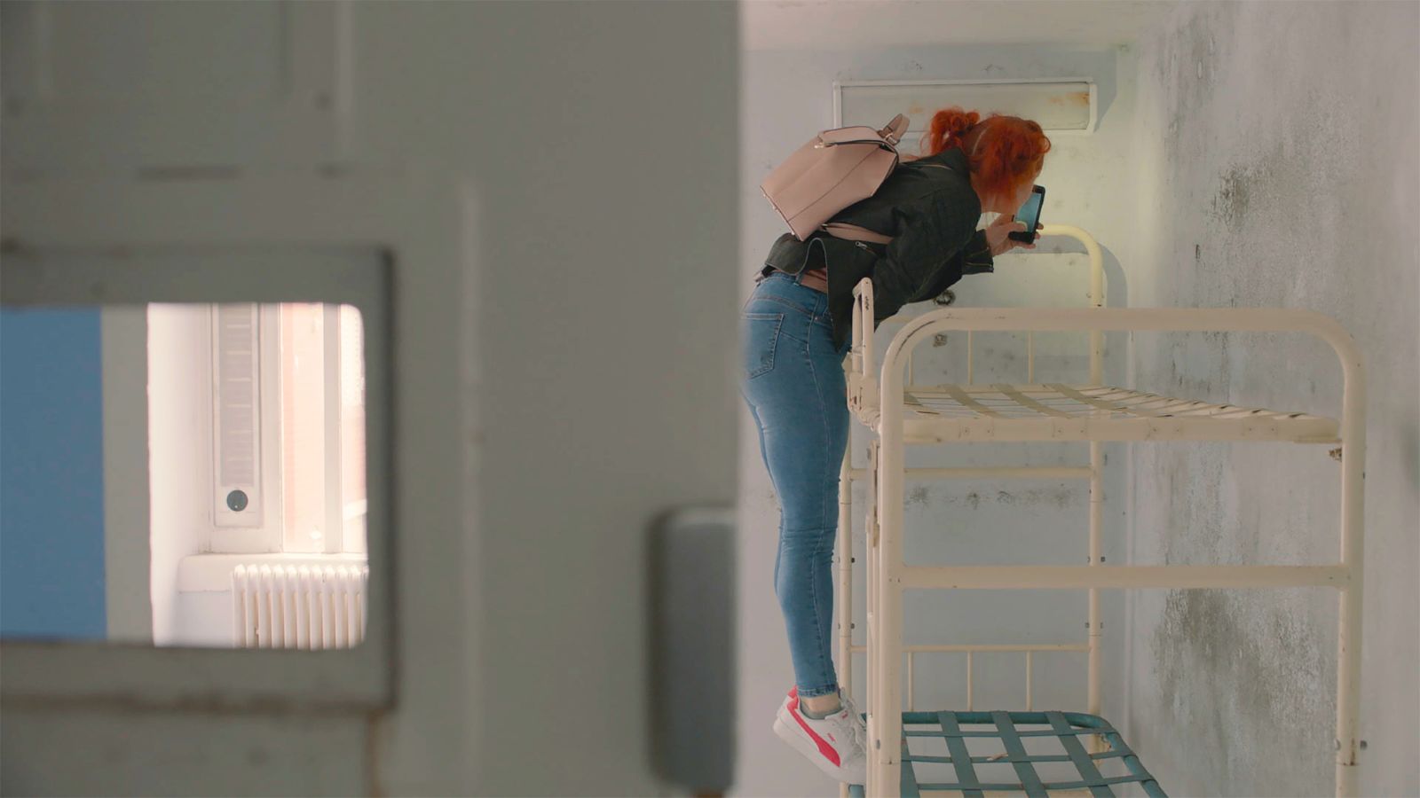 A woman wearing a black sweatshirt and jeans stands on the bottom bunk of a bunk bed and leans up onto the top bunk.