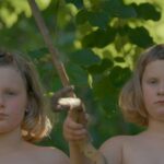 Two young sisters stand in front of the forest. They are both white with blonde hair, and pictured from the shoulders up.