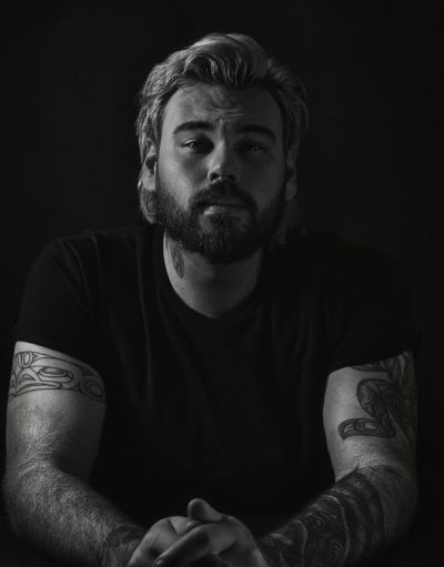 A black and white portrait of filmmaker Patrick Shannon. He is a Haida man with a beard and is wearing a black t-shirt with his arms displaying tattoo.s