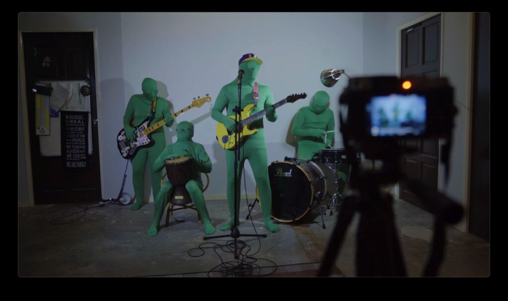 A punk band of four members performs on stage while a camera is in the foreground filming them. The musicians are dressed completely in green suits with their bodies and faces fully covered.