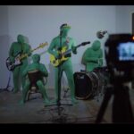 A punk band of four members performs on stage while a camera is in the foreground filming them. The musicians are dressed completely in green suits with their bodies and faces fully covered.